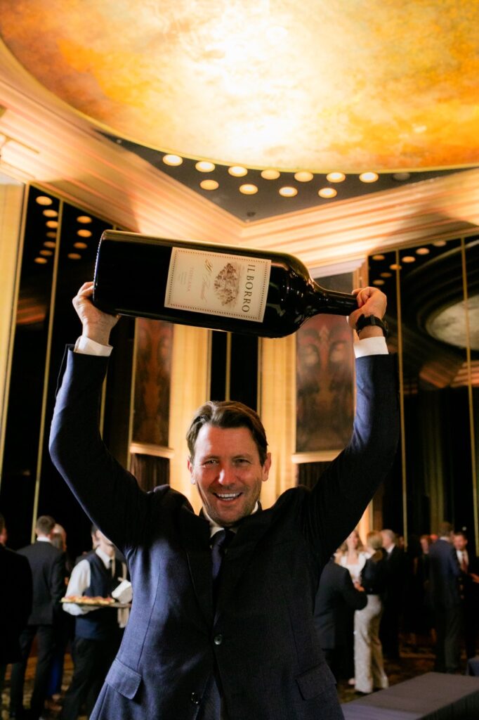 A man in a blue suit holds a very large bottle of Il Borro wine over his head.