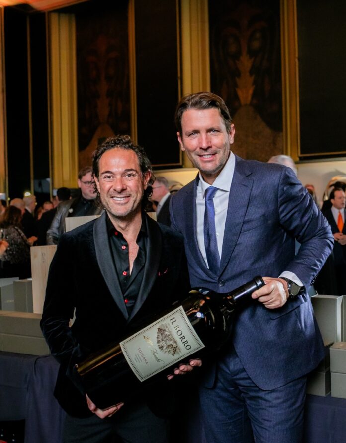 A man in a blue suit smiles next to a man in a black suit who is holding an obscenely large bottle of Il Borro wine.