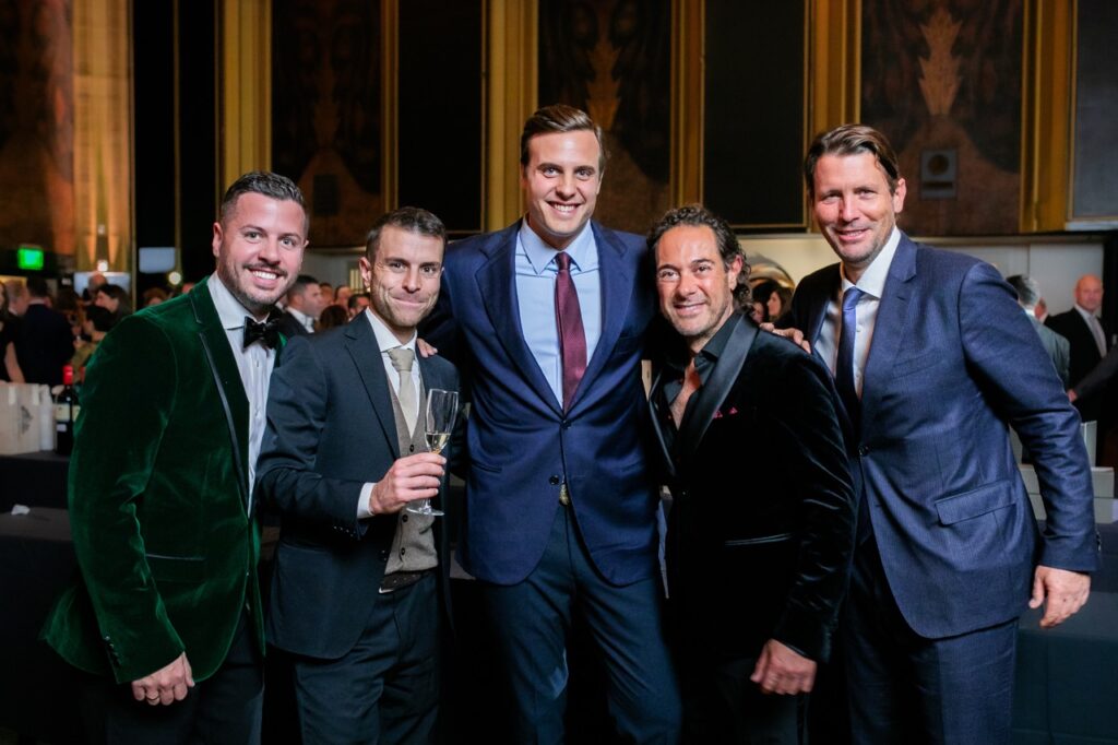 A group of men, two in blue suits, two in black suits, and one in a green suit jacket stand together and smile.