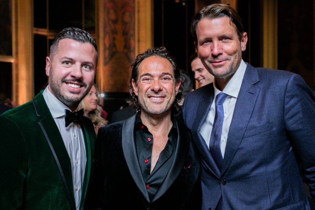 Three men in a green jacket, black jacket, and blue jacket stand together, smiling.