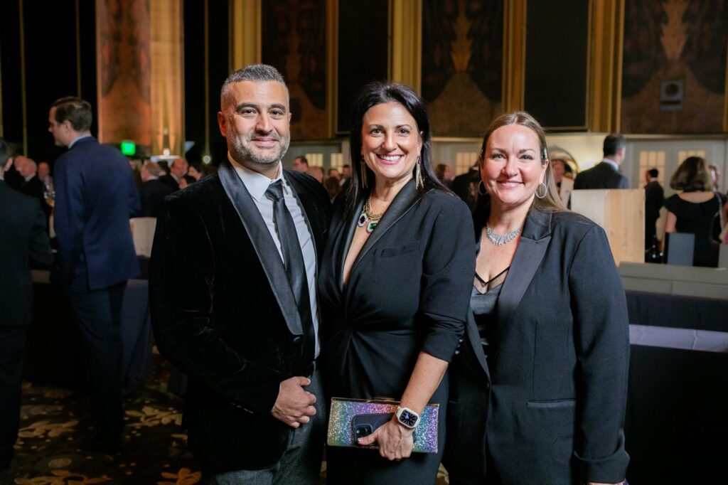 A man in a black suit jacket and tie stands next to a woman in a black suit jacket and another woman beside her in a black suit jacket.