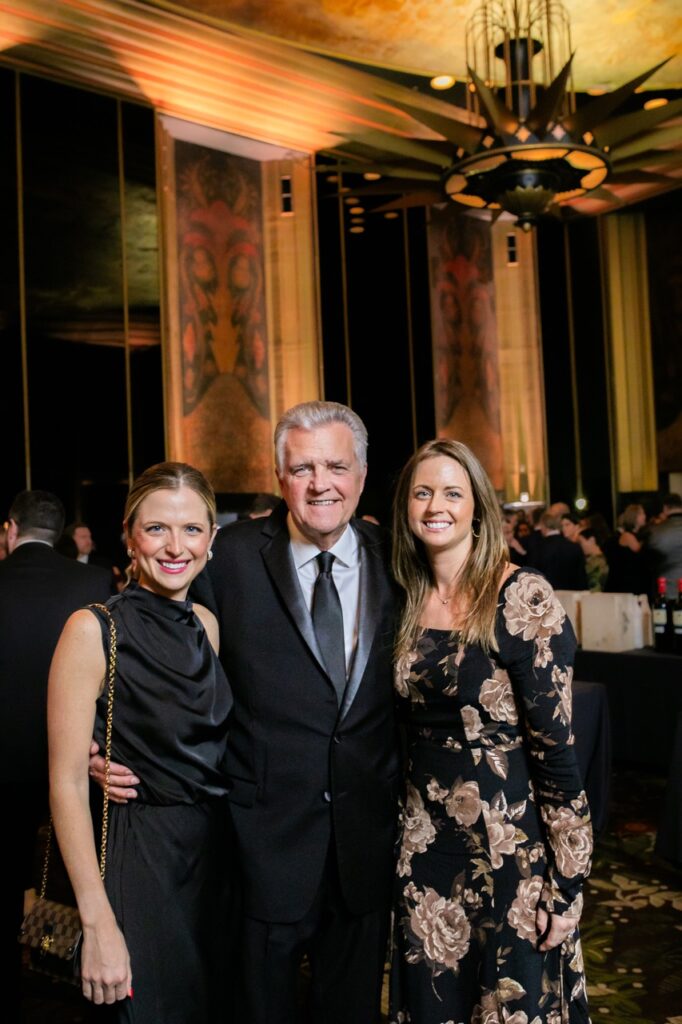 Two women in dresses stand on both sides of an older man in a black suit and tie.