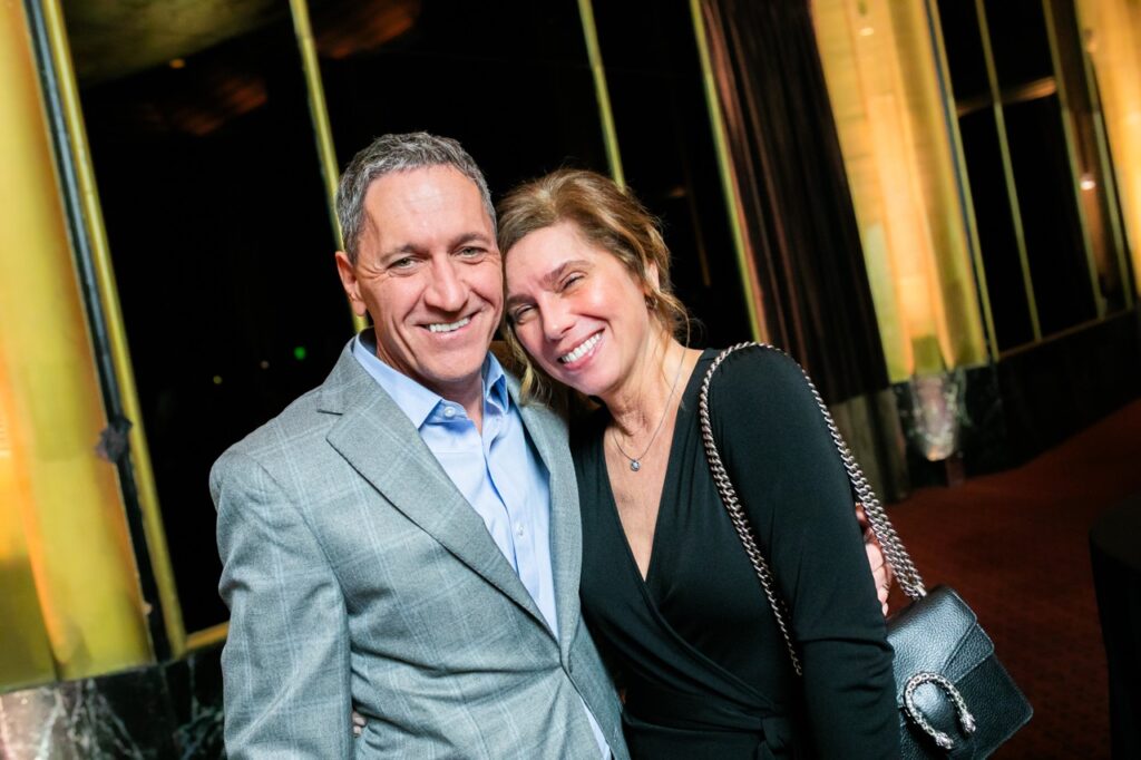 A woman in a black dress smiles with a man in a grey, plaid style suit jacket and blue undershirt.