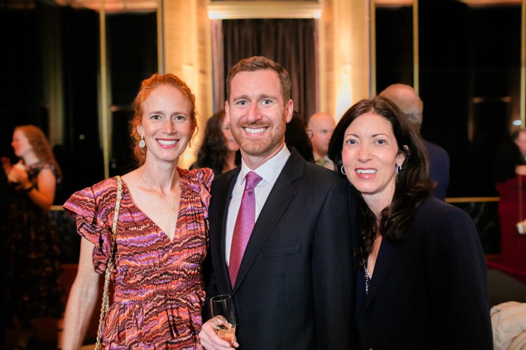 A woman in a colorful red and purple dress stands with a man in a black jacket holding a champagne glass and a woman in a black suit jacket.
