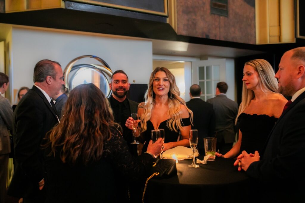 A woman in a black dress smiles and holds a champagne glass amongst other persons in black dress clothes.