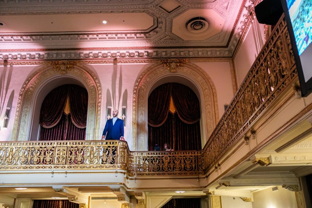 A man in a blue suit sings from the balcony of a high class regency room.