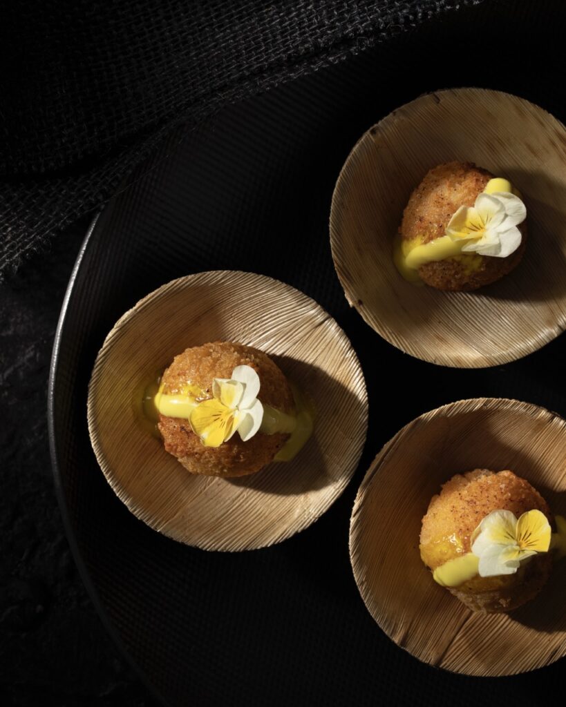 Three brown wood bowls hold lumps of cake cakes with a yellow sauce over top and white and yellow edible flowers.