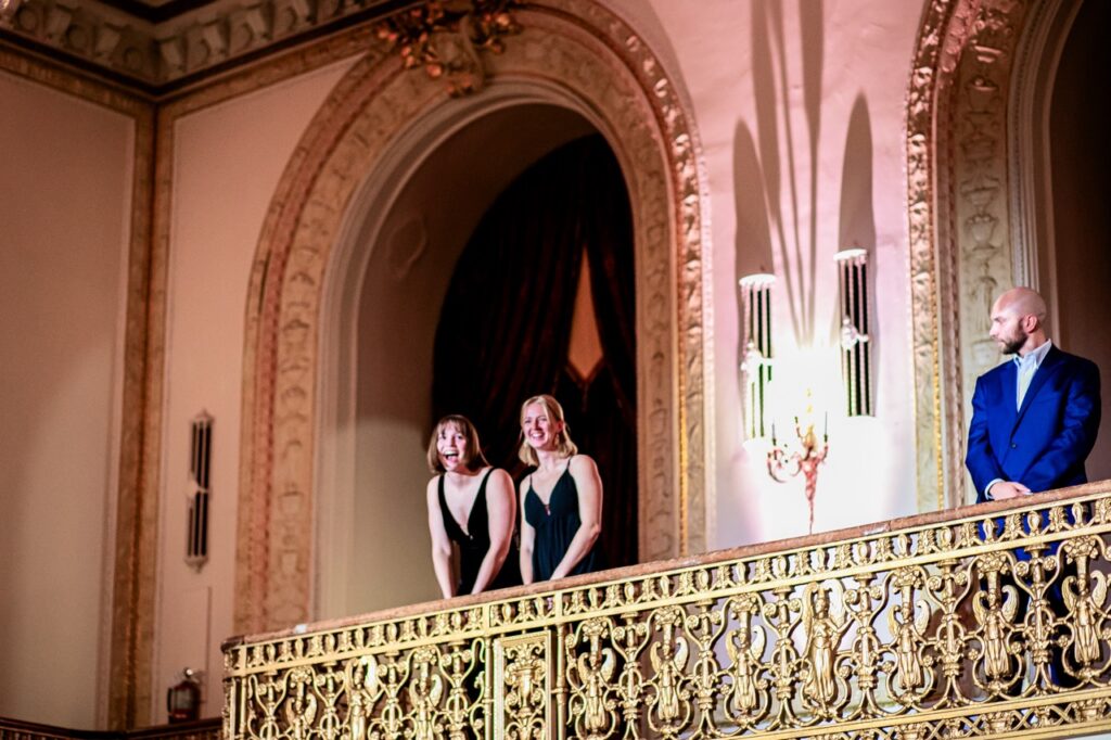 Two women in black dresses smile and sing from a gold balcony as a man in a blue suit watches.