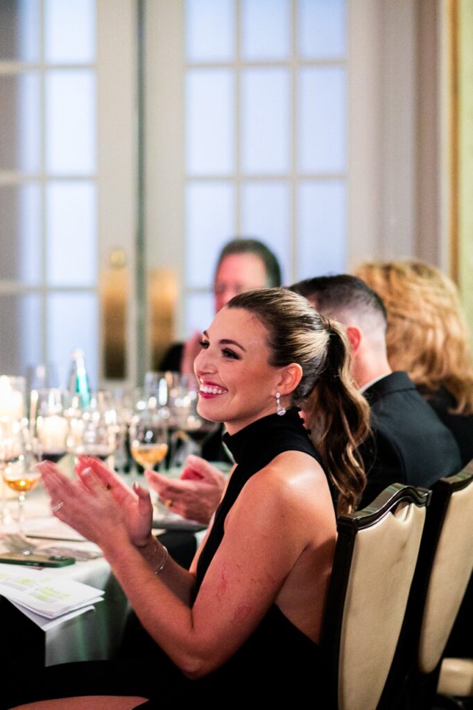 A woman in a black dress with her hair in a ponytail sits at a table, clapping her hands.