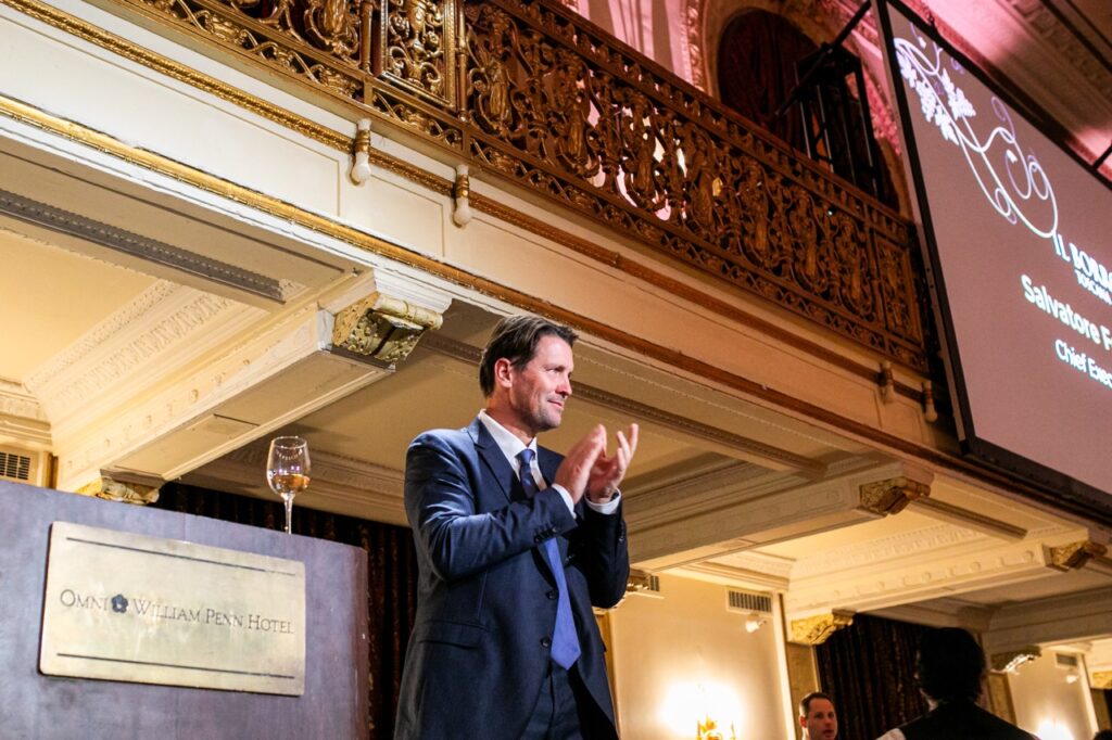 A man in a dark blue suit claps his hands under a gold balcony and screen.