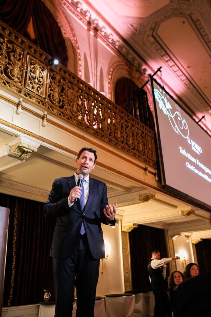 A man in a dark blue suit uses a microphone as he talks to a room full of people.
