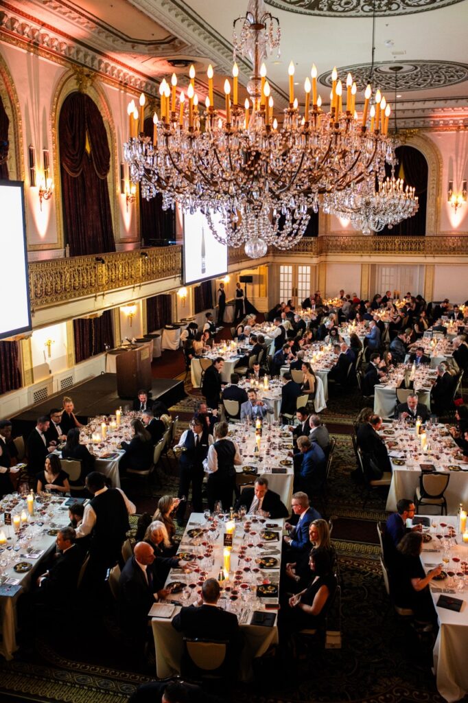 A bunch of rectangular tables with people sitting at them sit underneath a huge chandelier on the ceiling.