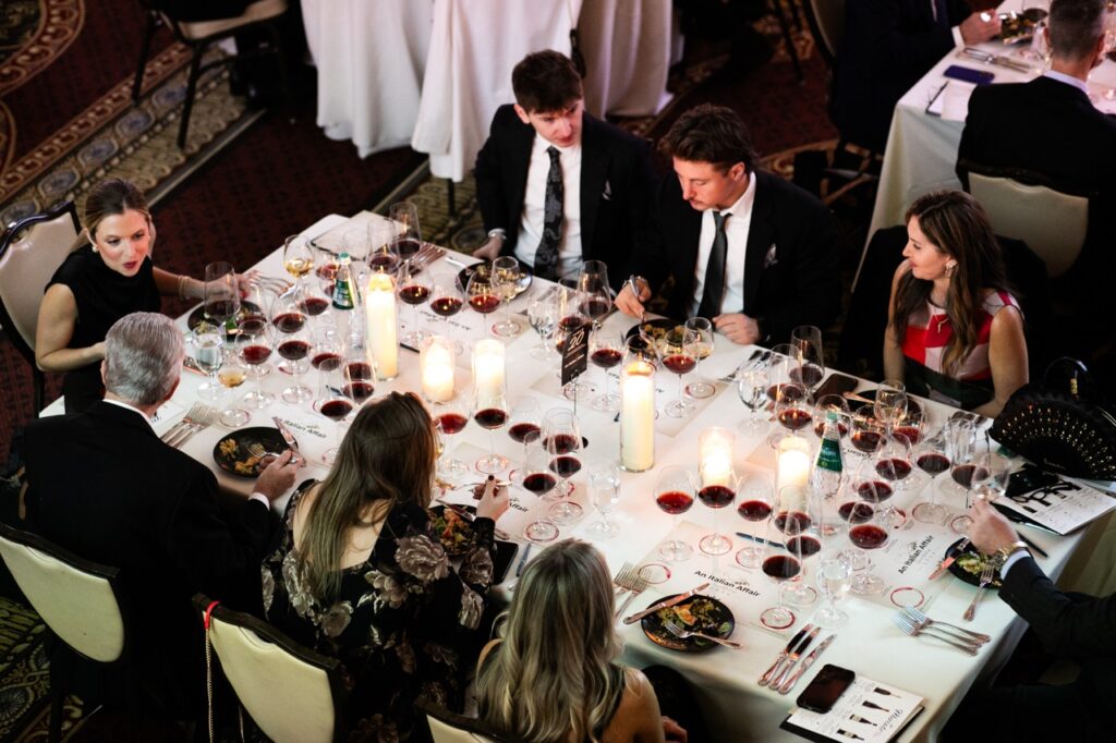 A group of 8 people sit at a white rectangular table with wine glasses and plates of food scattered throughout.