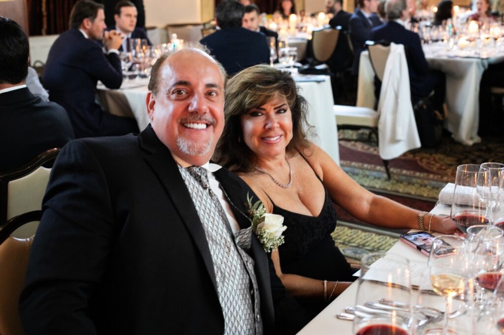 A man in a black suit jacket with a white flower pinned to it smiles with a woman in a black dress.