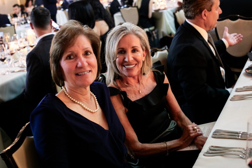 Two women, one in a blue dress with pearls, and another in a black dress with a simple gold chain, smile together.