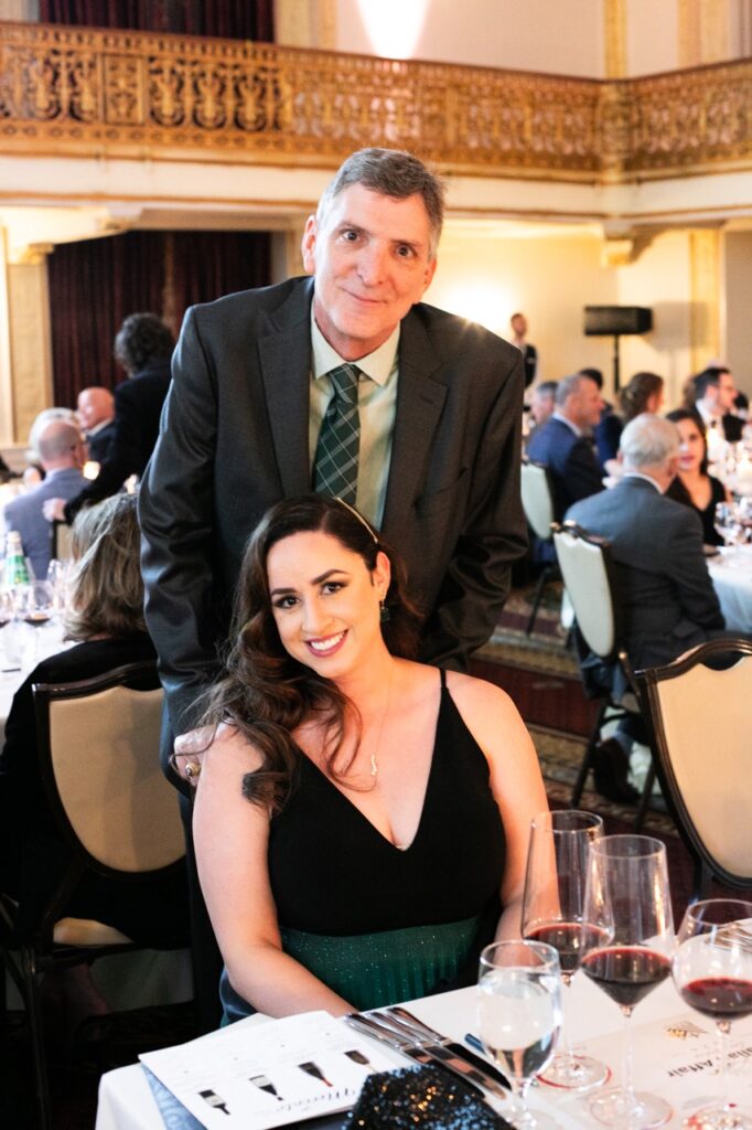 A man in a grey suit jacket stands behind a woman in a black and green dress who is sitting in a chair.