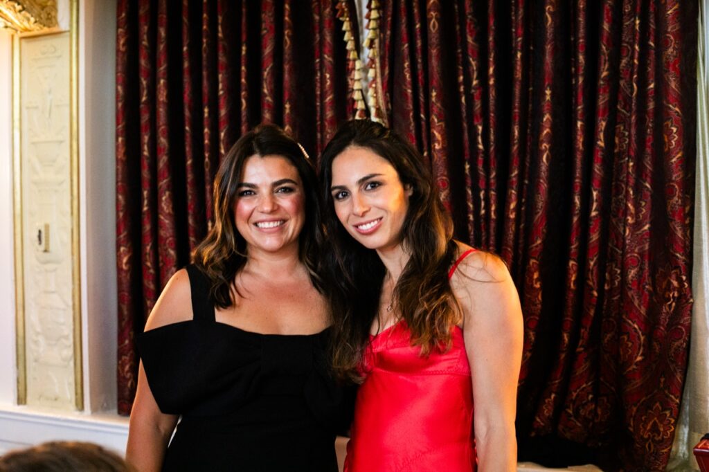 A woman in a red dress and a woman in a black dress smile in front of a red print curtain.