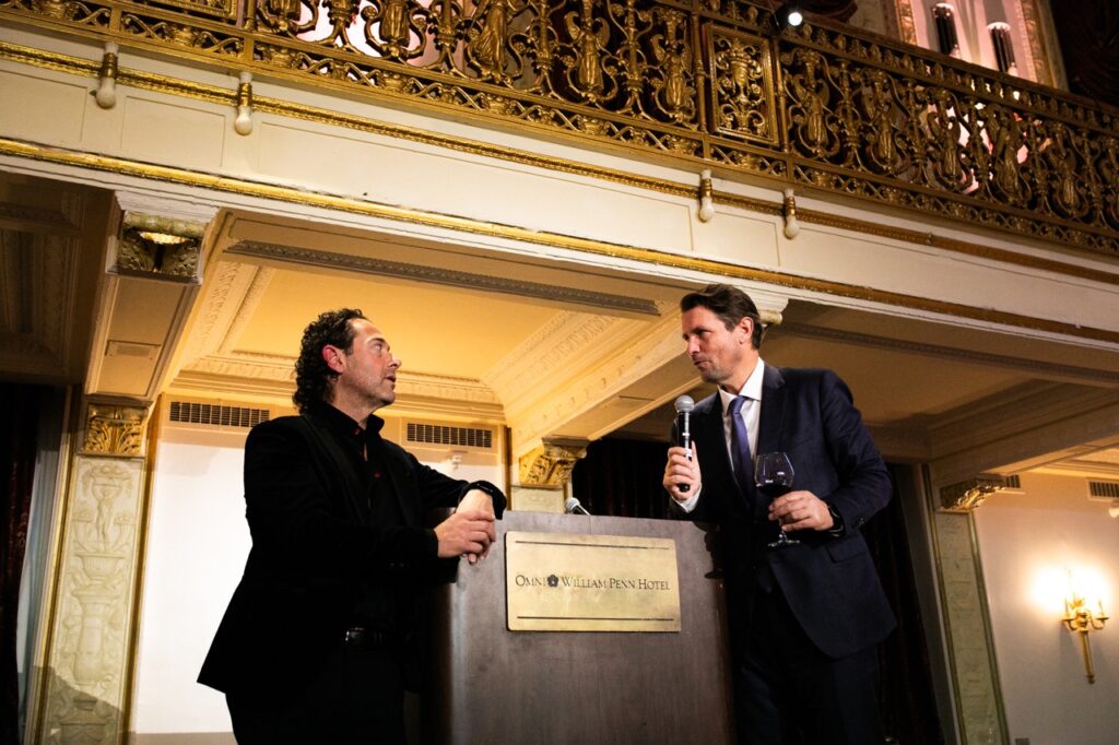 A man in a black suit and a man in a blue suit with a glass of wine and a microphone speak to one another over a podium.