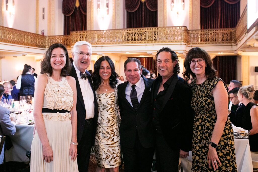 A group of three men in black suit jackets and three women in dresses stand together in a ballroom.
