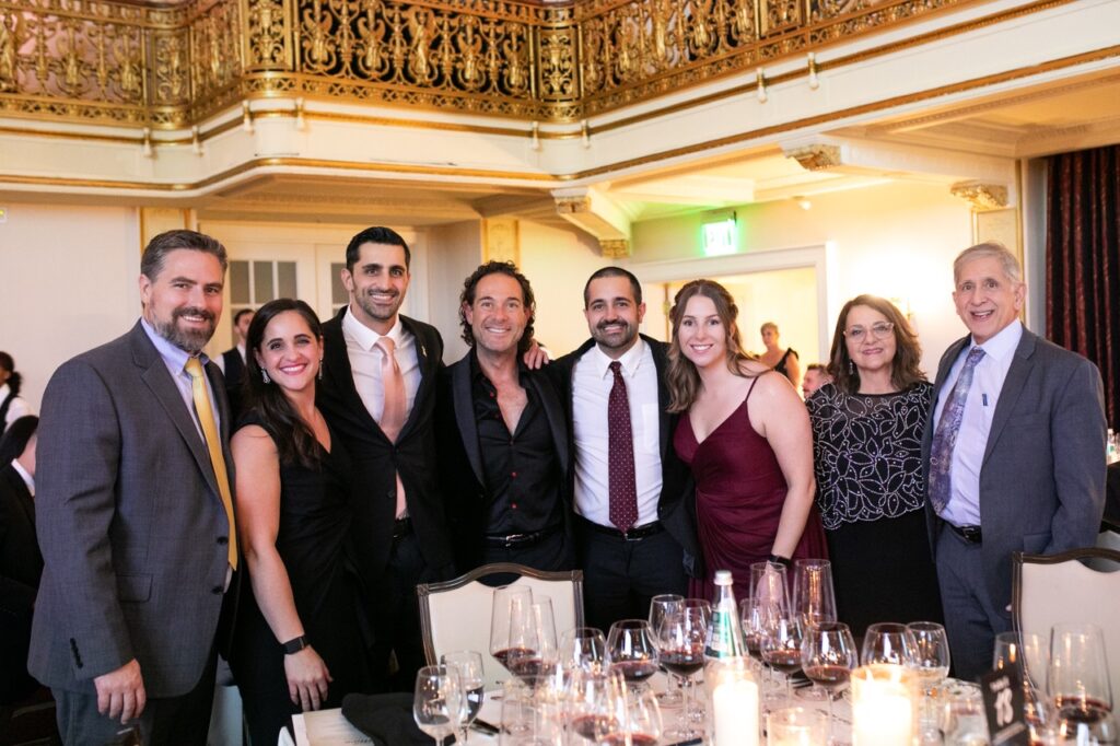 A group of three women in dresses and five men in suit jackets stand arm and arm behind a table scattered with wine glasses.