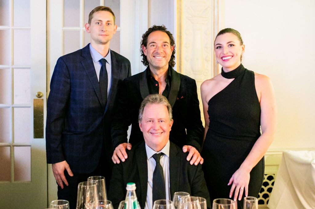 A man in a black suit jacket stands with his hands on a man's shoulders sitting in front of him with a woman on his right and another man on his left.