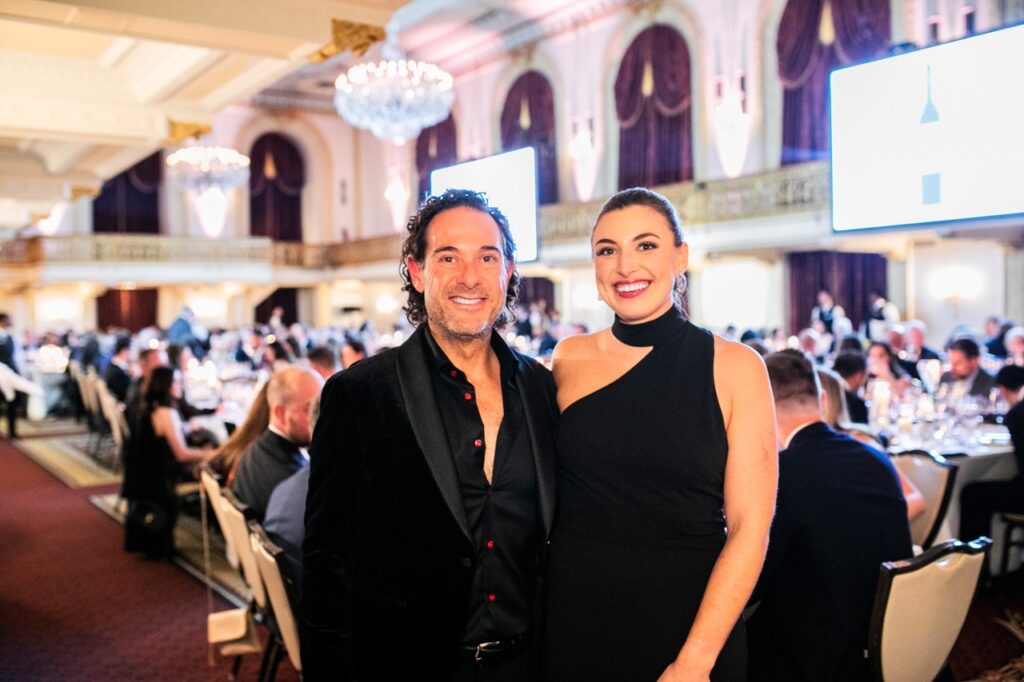 A man in a black suit and a woman in a black dress smile in front of a huge room full of people sitting down at tables.