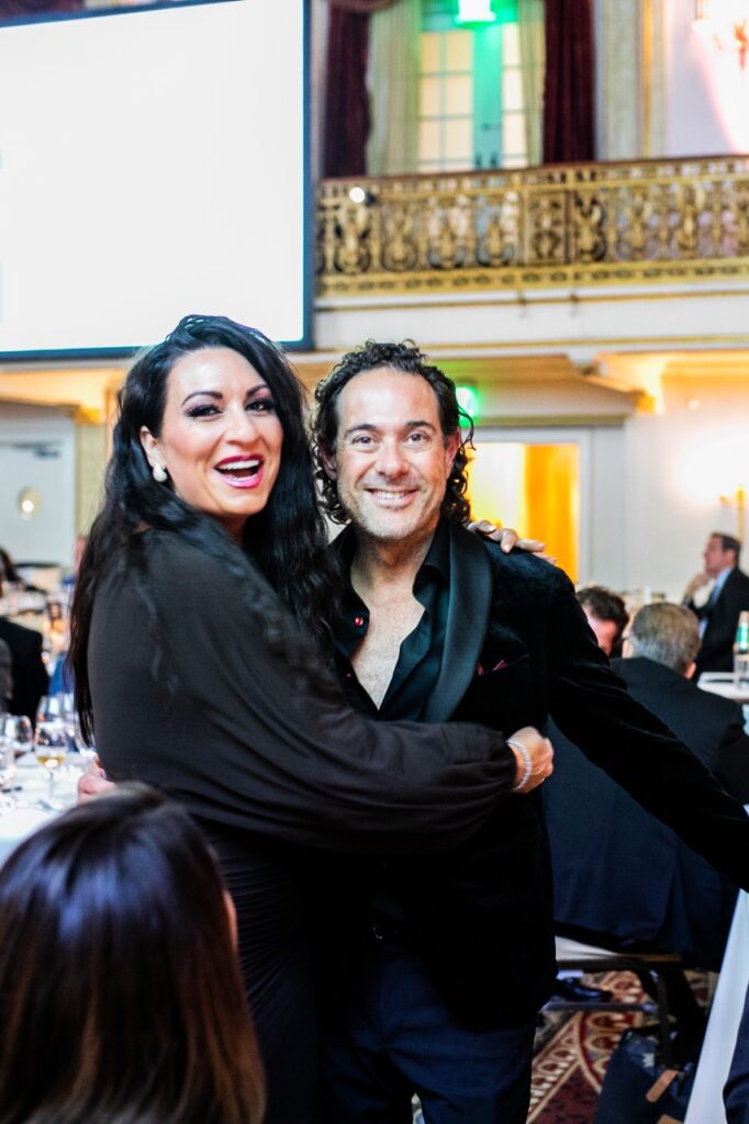 A woman in black hugs on to a man in a black suit jacket.