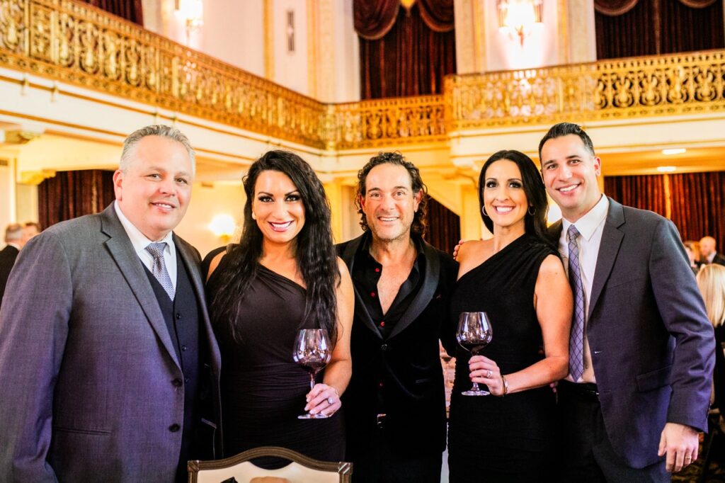 Two women in black dresses with wine glasses in their left hand stand with three men in suit jackets.