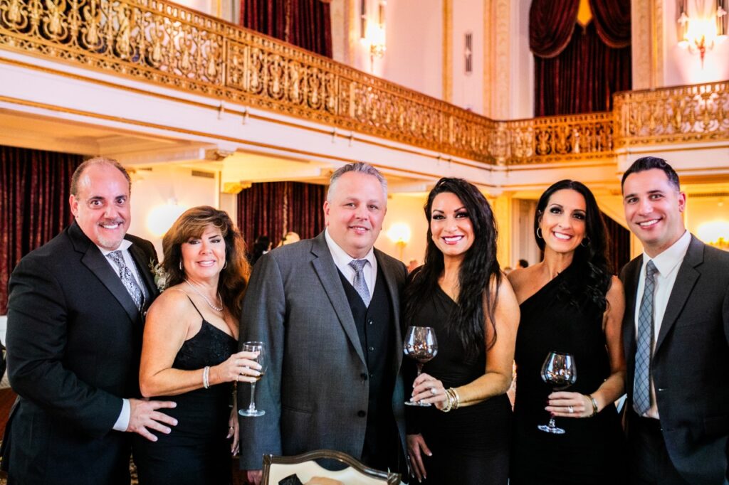 Three women in black dresses, each holding a wine glass stand with three men in different colored suit jackets.
