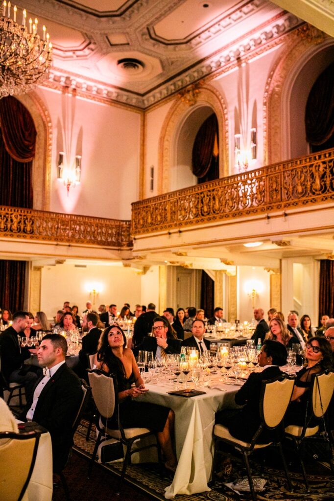 People sitting at tables with wine glasses and candles look up towards the ceiling of a ballroom.