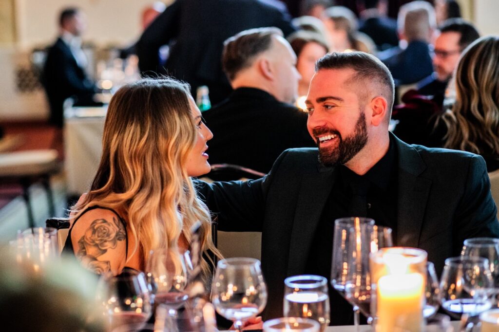 A man in a black suit jacket smiles and talks to a woman in a black dress with blonde hair and a tattoo.