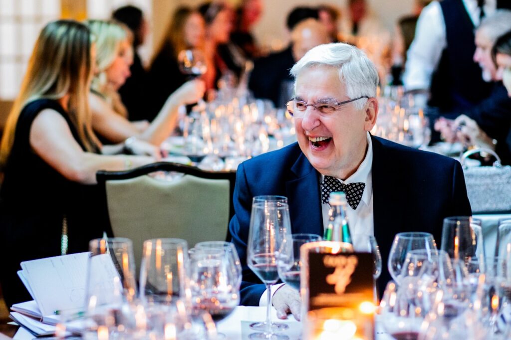 A man in a dark blue suit jacket and bowtie with glasses laughs hard.