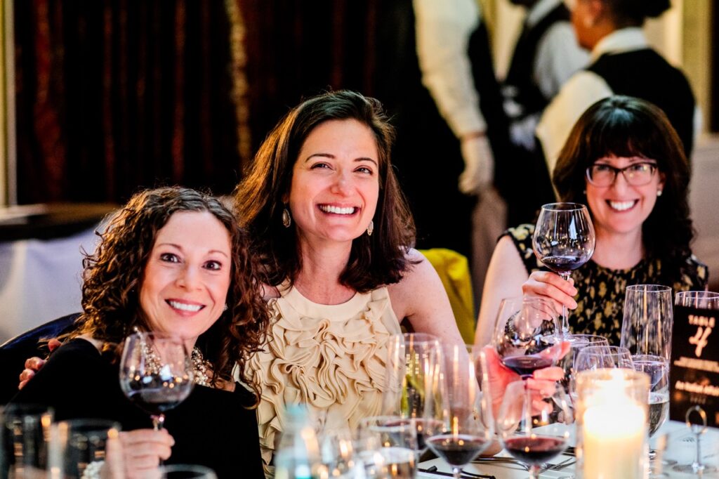 Three women smile while sitting at a table, wine glasses in hand.