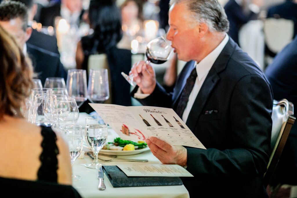 A man in a black suit jacket looks at a folder full of wine options as he sips from a wine glass.