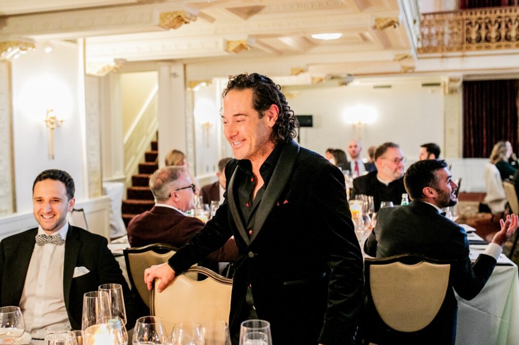 A man in a black suit jacket stands with his hand on a beige chair next to a man sitting with a bowtie on.