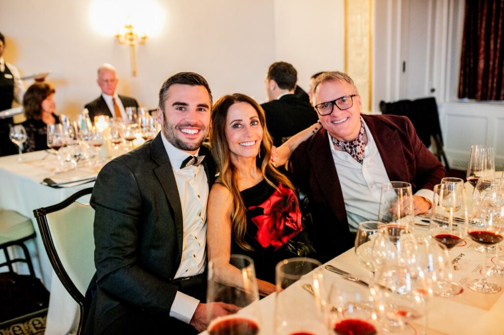 A man in a black suit jacket and bowtie sits with a woman in a black and red dress leaning back against him with her arm around a man in a red suit jacket.