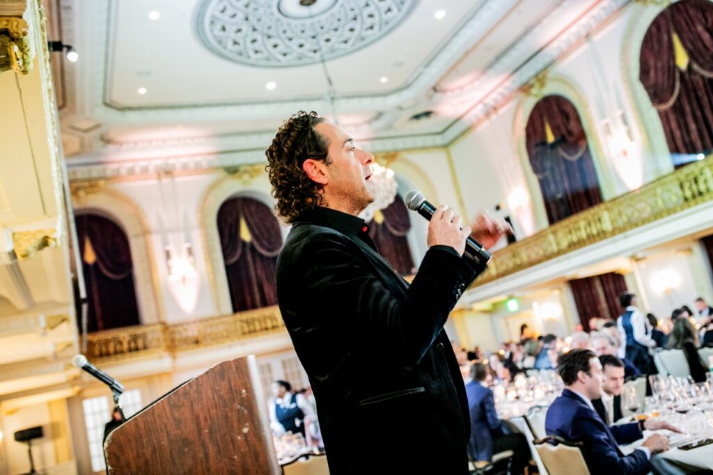 A side profile of a man in a black suit jacket as he speaks into a mic on a stage.