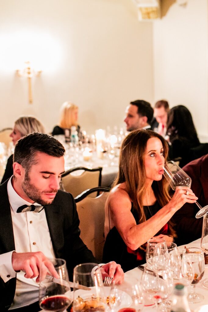 A woman tastes from a large wine glass while a man cuts his meal at the table.