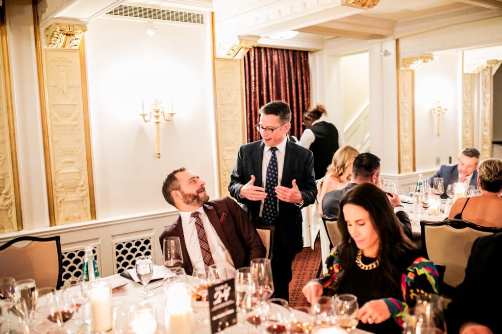 A man in a black suit jacket and tie talks to a man in a red suit jacket sitting at a table while a woman beside them cuts her meal on her plate.