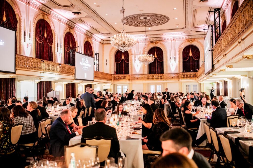 A large ballroom with chandeliers holds many people sitting at large rectangular tables filled with wine glasses.