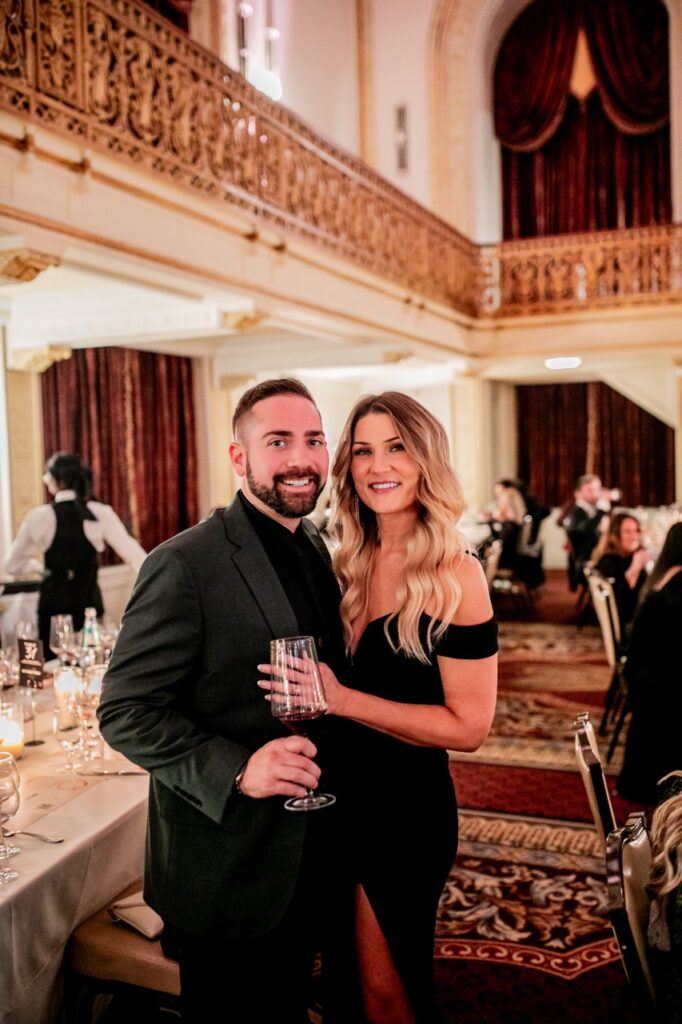 A man in a black suit jacket and glass of wine in hand poses with a woman in a black dress who has her hand on his jacket.