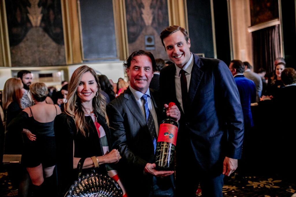 A man in a grey suit jacket and blue tie holds a large bottle of wine in between a woman in a black dress and a man in a dark blue suit jacket.