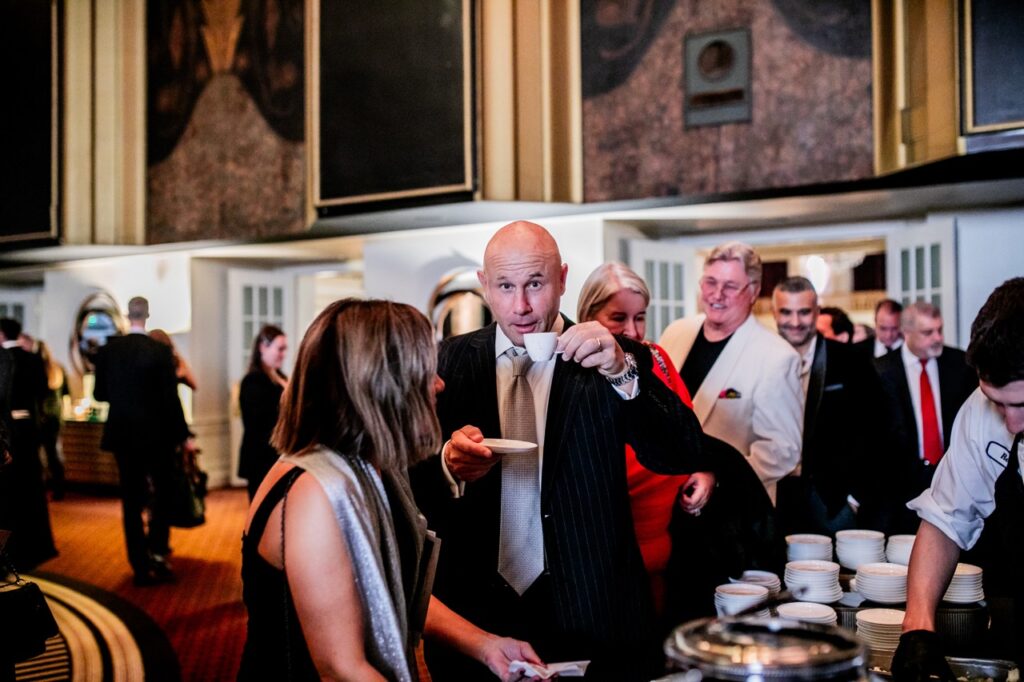A man in a black suit jacket holds up a small cup of coffee with a small plate in his other hand.