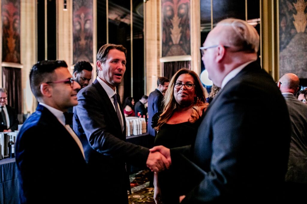 A man in a blue suit shakes hands with a man in a black suit as another man and woman watch the two.