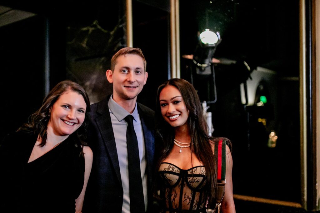 Two women stand with a man in a suit jacket and tie between them.