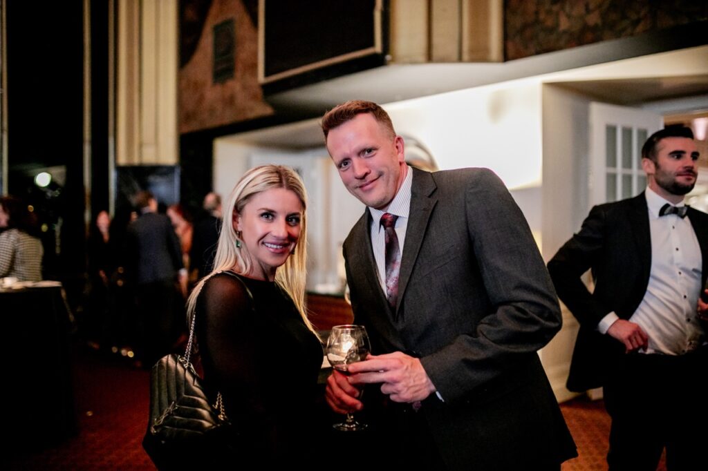 A man in a grey suit jacket with a glass of wine leans down to smile with a woman in a black dress.