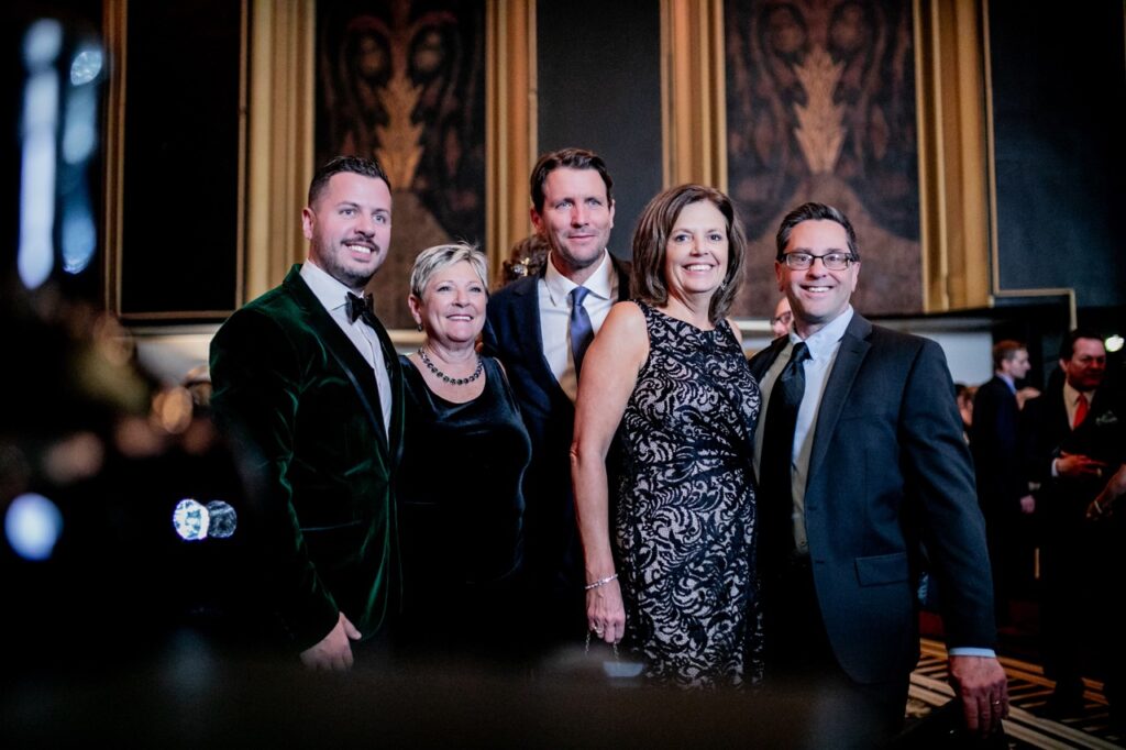 A man in a green suit jacket smiles with a man in a black dress, a man in a blue suit, another woman in a white and black dress, and a man in a dark blue suit.