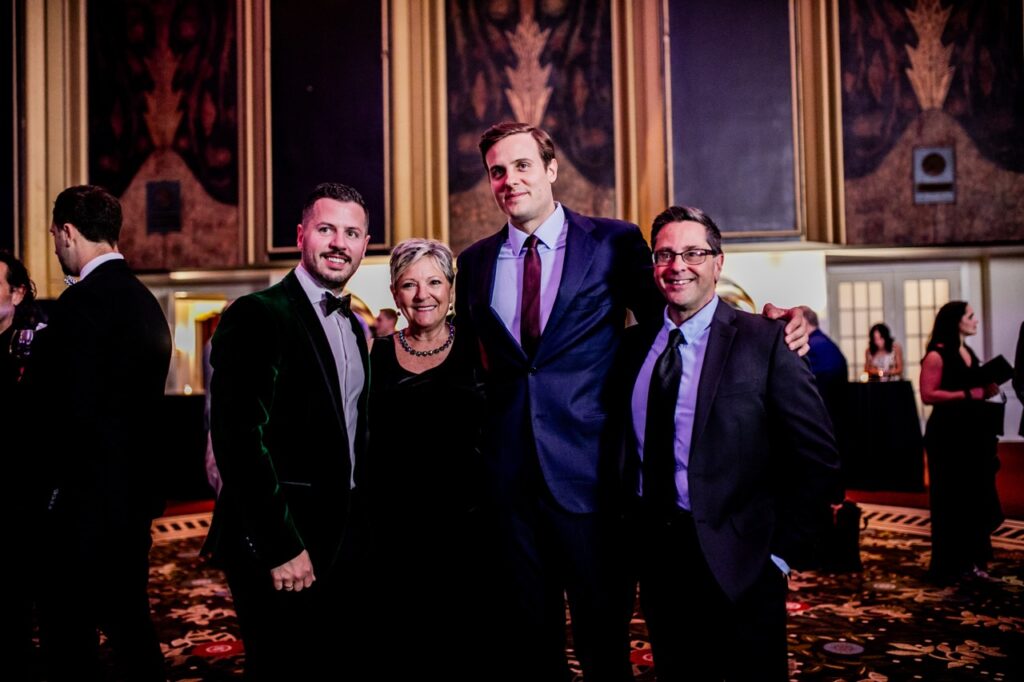 A man in a green suit stands with a woman in black, and two other men in blue suits.