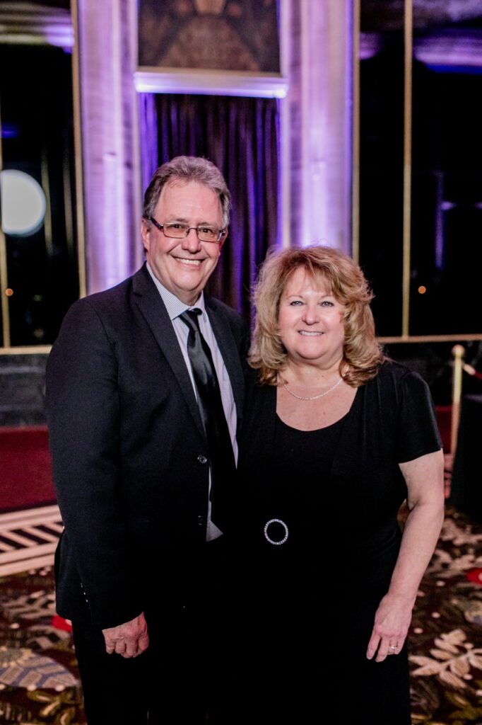 A man in a black suit jacket and tie smiles with a woman in all black.