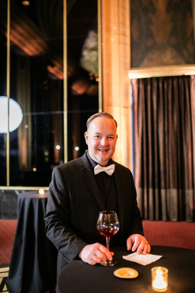 A man in a black suit jacket and white bowtie holds a glass of wine on a black table by the bottom of its stem.